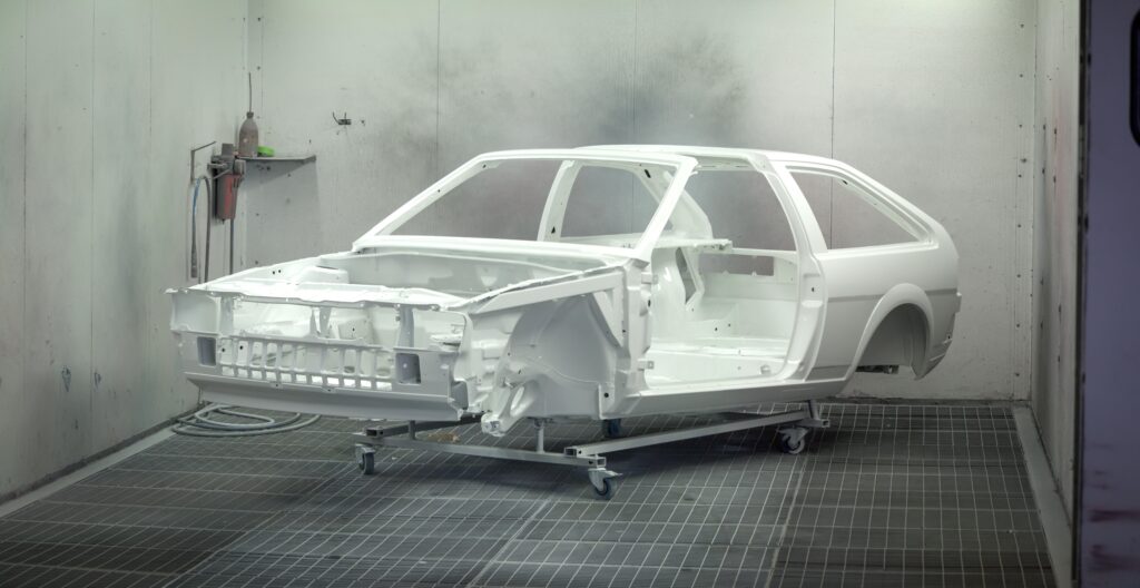 A vintage car frame being painted white in an industrial automotive paint booth.