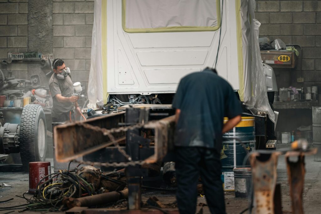 Two workers in a workshop painting a vehicle chassis. Industrial setting, protective gear.