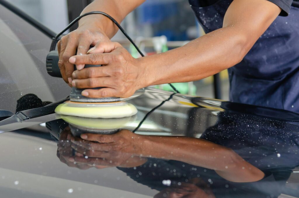 Detailed view of a worker's hands polishing a car's hood using a buffer in an auto workshop.