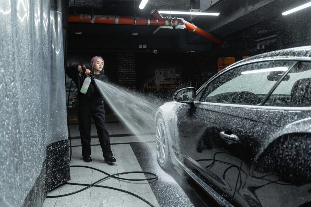 Woman using a pressure washer to clean a black car in an indoor car wash setting.