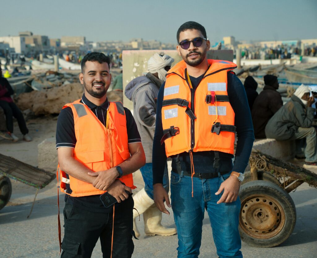 Smiling men standing in life vests at a bustling dock area under bright sunlight.